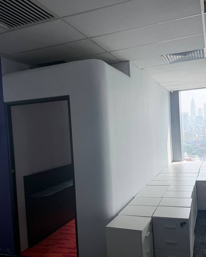 Curved white plaster partition wall with rounded corner beneath a suspended grid ceiling in Brickfields office, overlooking Kuala Lumpur skyline through window