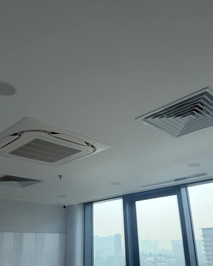 Flat plaster ceiling in Brickfields office with cassette air-conditioning unit, square diffuser vent, downlights and recessed smoke detector, floor-to-ceiling windows overlooking city skyline