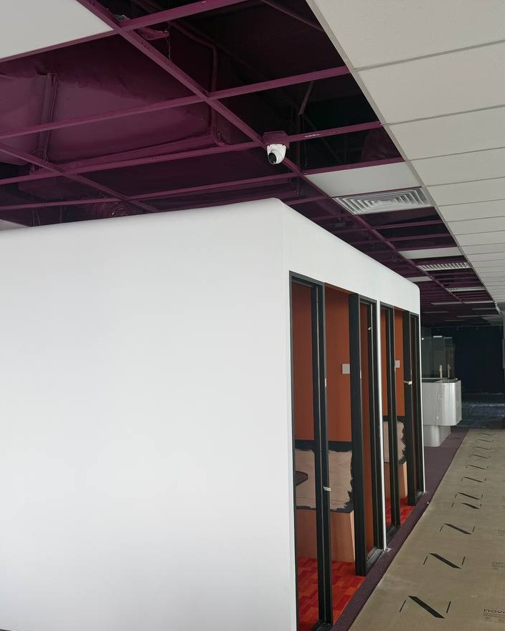 White partition wall corner with glass-panelled office pods and exposed purple ceiling structure at Brickfields office renovation
