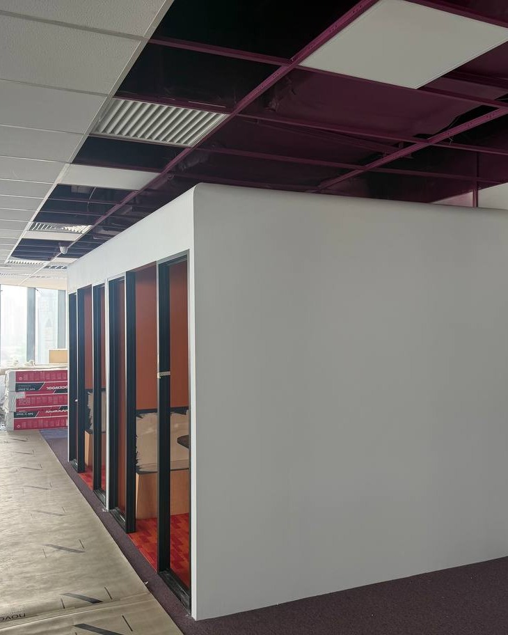 Freshly installed white partition wall enclosing a soundproof meeting room with black-framed glazed doors and red interior flooring, exposed maroon ceiling grid and suspended ceiling tiles overhead, Brickfields office fit-out