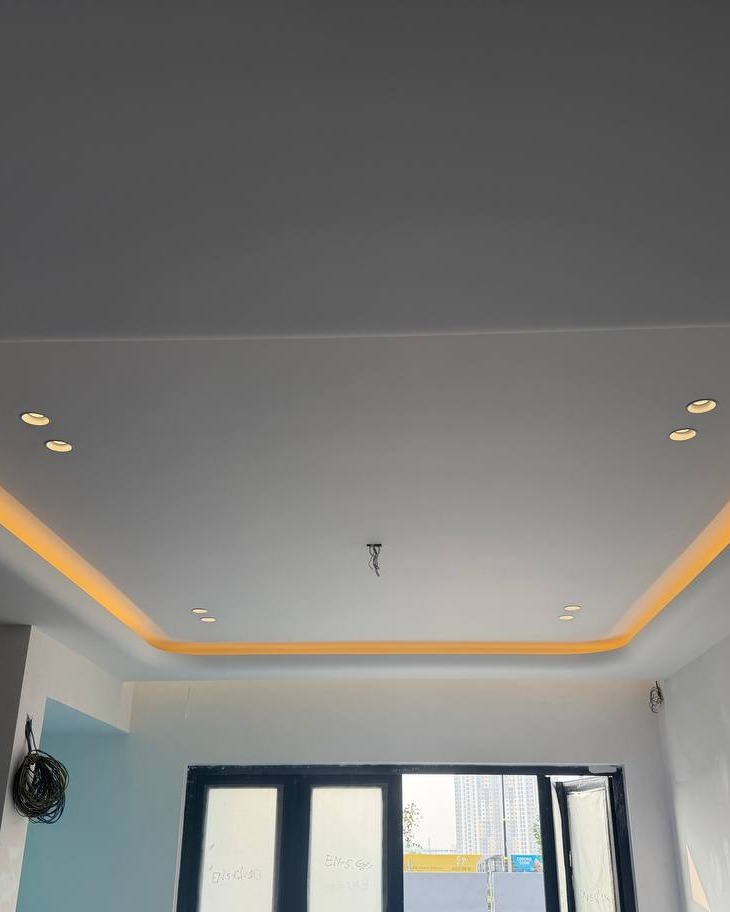 Recessed rectangular plaster ceiling drop with warm yellow LED cove lighting and multiple downlights, Klang residence installation, unfinished walls and pending window grille below
