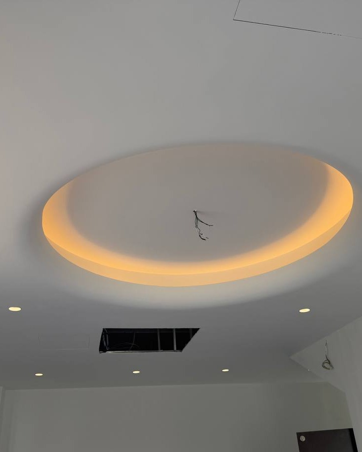 Oval plaster ceiling drop with recessed cove lighting trough glowing warm amber, Klang residence false ceiling installation mid-wiring stage