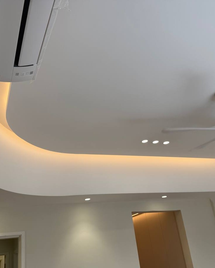 Curved plaster ceiling drop with warm LED strip lighting and recessed downlights, installed around a wall-mounted air conditioning unit in a Rawang home
